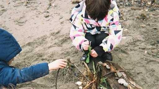 Two children in winter coats are playing on the beach with natural found items.