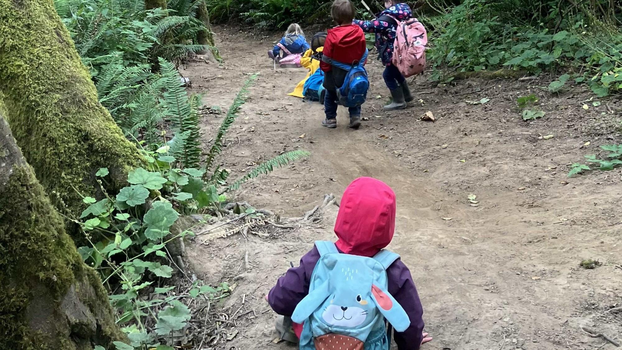 Four children walk on a dirt trail with one carrying a blue bunny backpack.