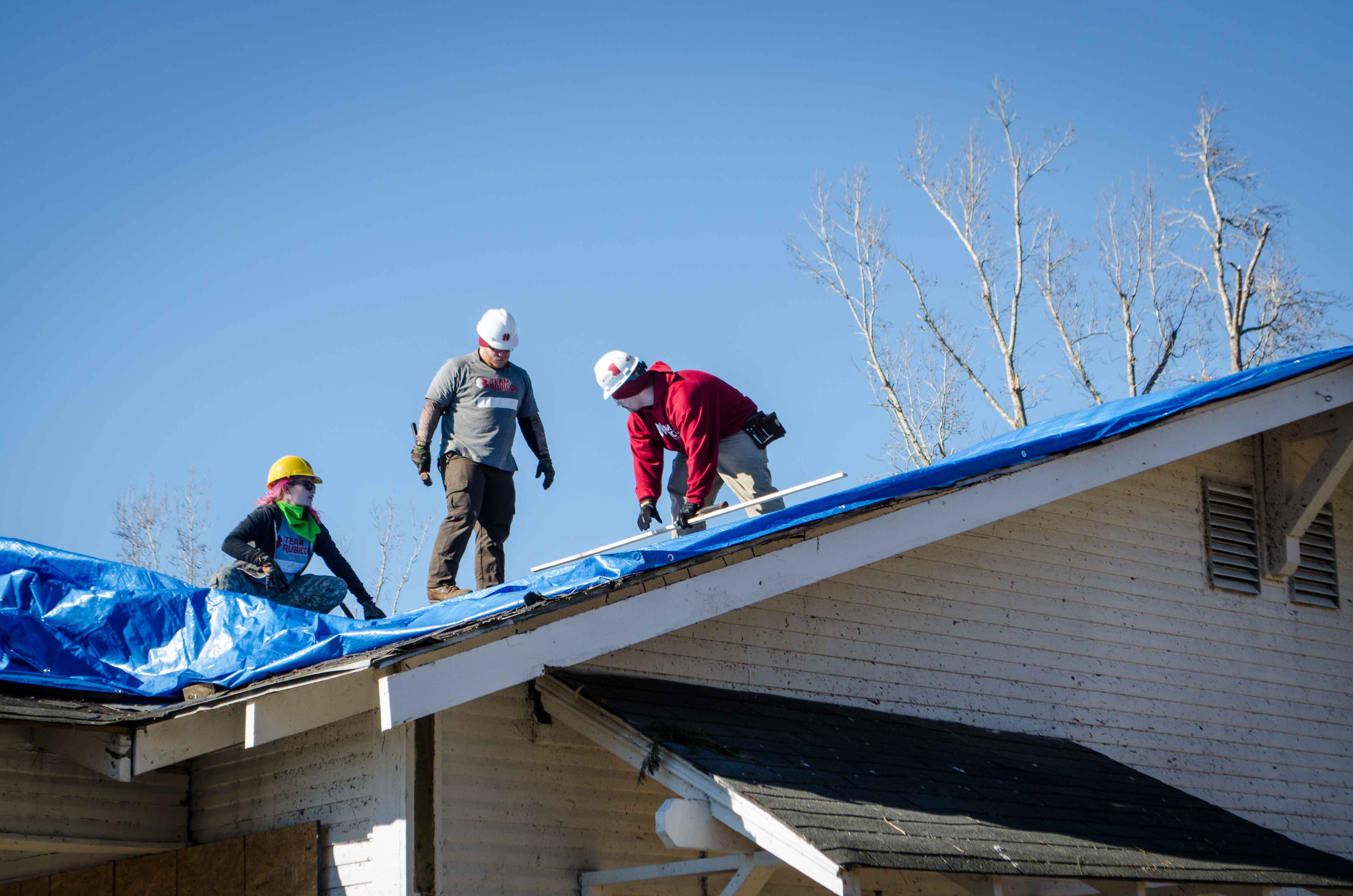 Three Greyshirt volunteers stand on a roof. They are actively putting on a blue tarp, shielding the home from further water damage.