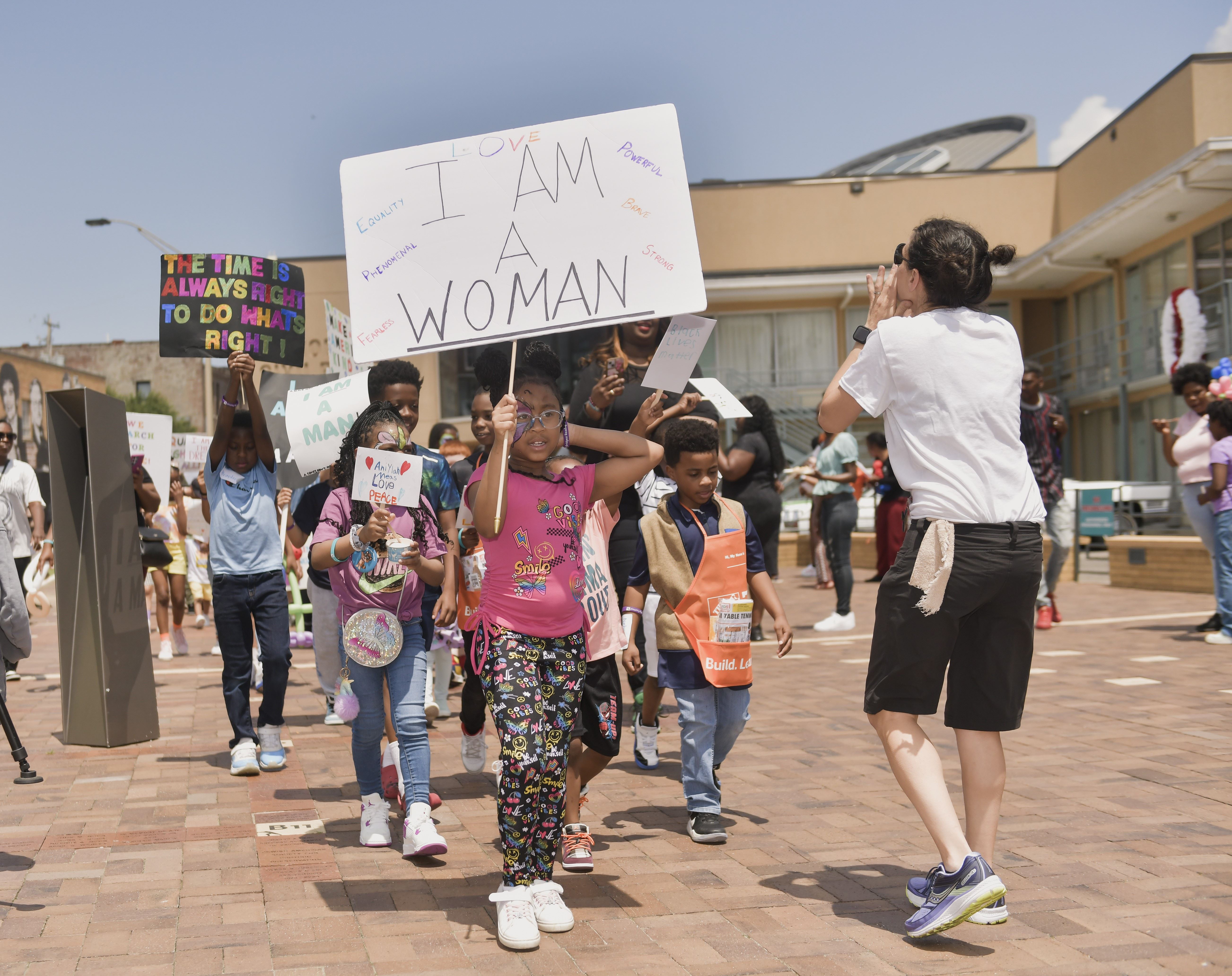 Students march at the Ruby Bridges Reading Festival, led by girl with an I Am A Woman sign