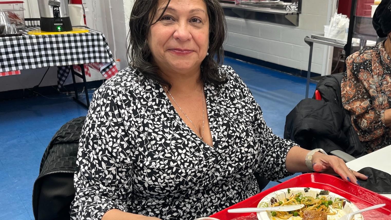 A woman sits at a table in a community center eating a healthy lunch off a bright red cafeteria tray.
