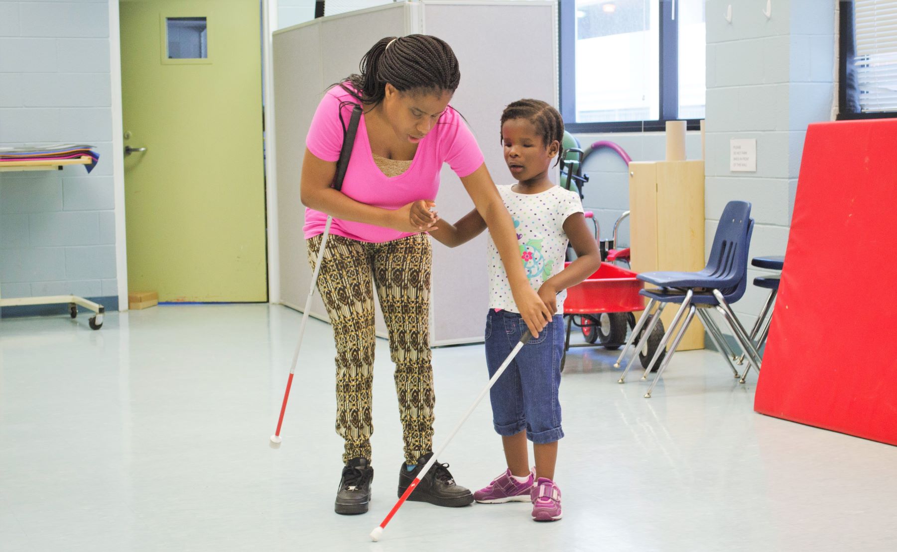 An adult holding a white cane teaches a young girl how to hold a white cane in a spacious room with chairs and gym mats in the background.