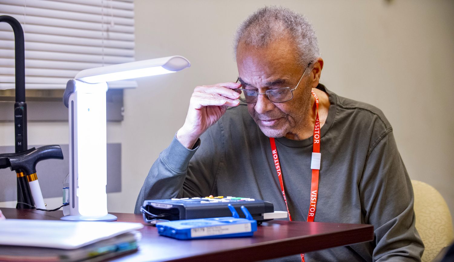A man sitting at a table using a bright table lamp to read a book.