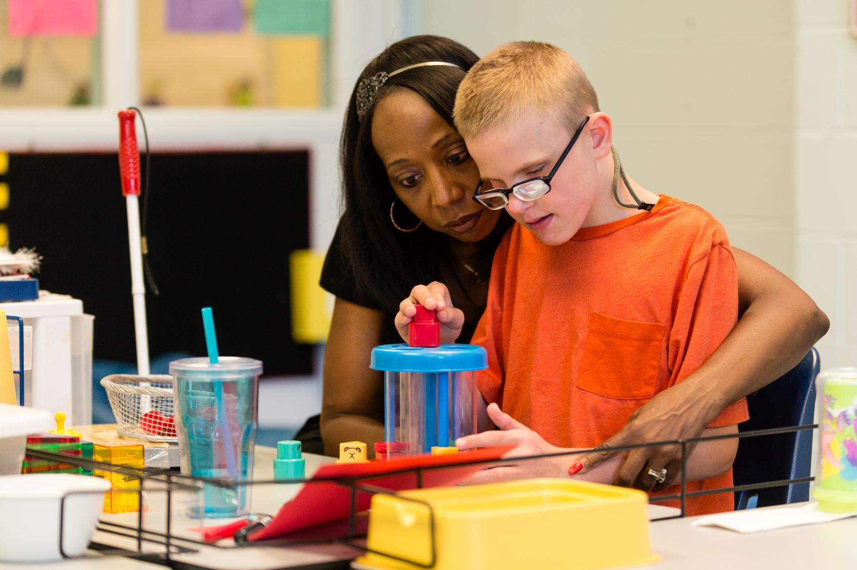 A teacher assisting a student with an educational activity at a classroom table filled with colorful learning materials.