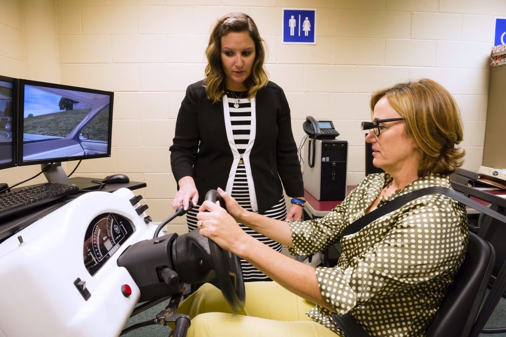 A woman sits in a driving simulator with bioptic telescopic glasses on, which look like regular glasses but with a small telescope coming out of the center.