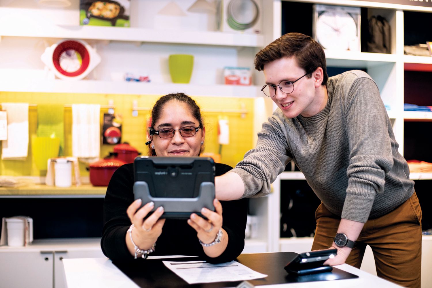 A man is instructing a woman on how to use a handheld digital device that she is holding up in front of her face. They are in a bright, colorful store.