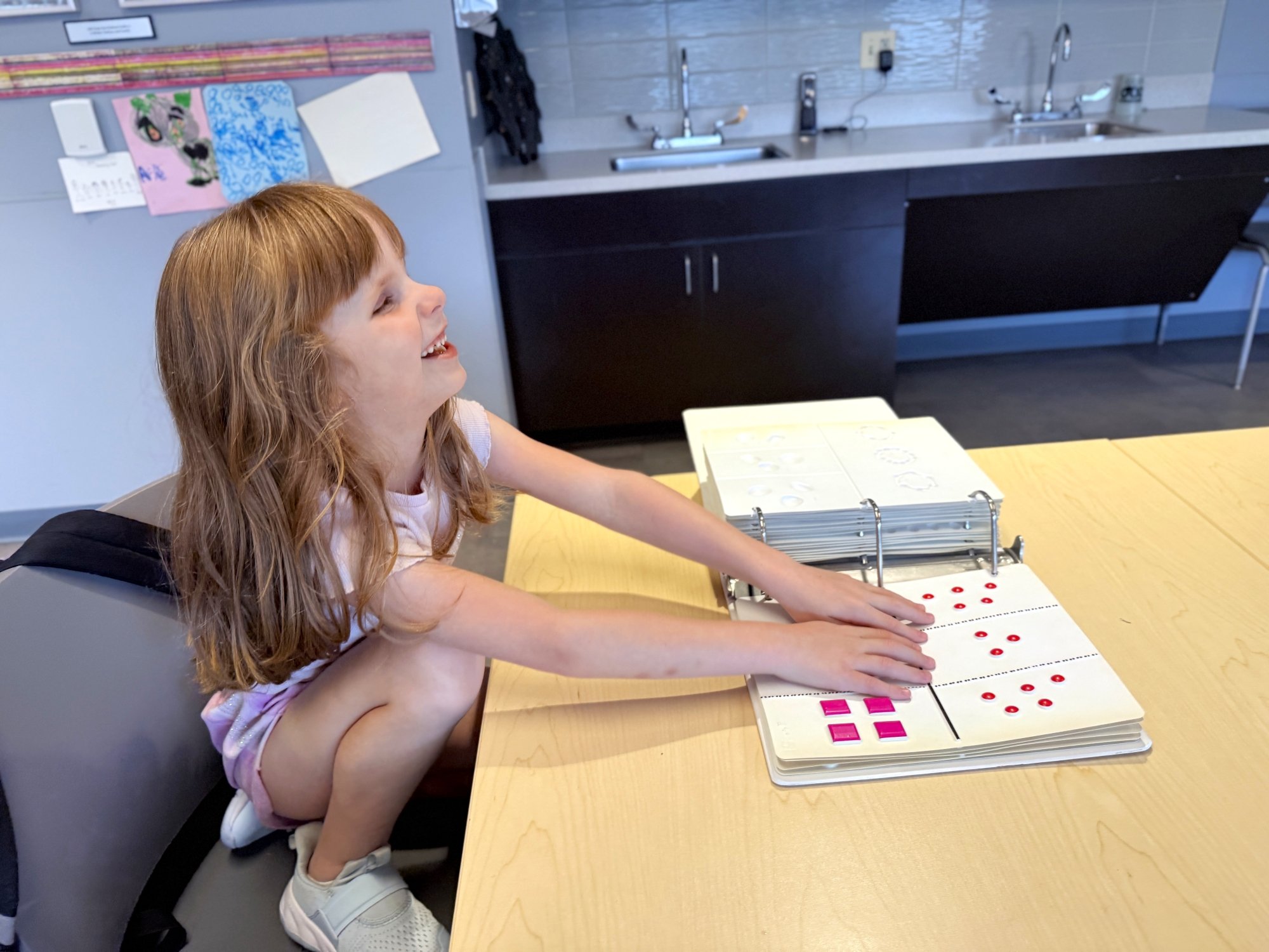 A young white girl smiles as she feels the letters in a Braille reader.