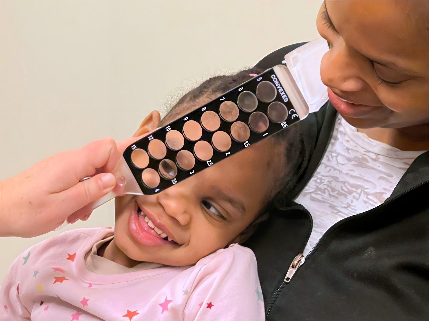A child smiles while receiving an eye exam with an eye occlusion paddle, held by an adult.