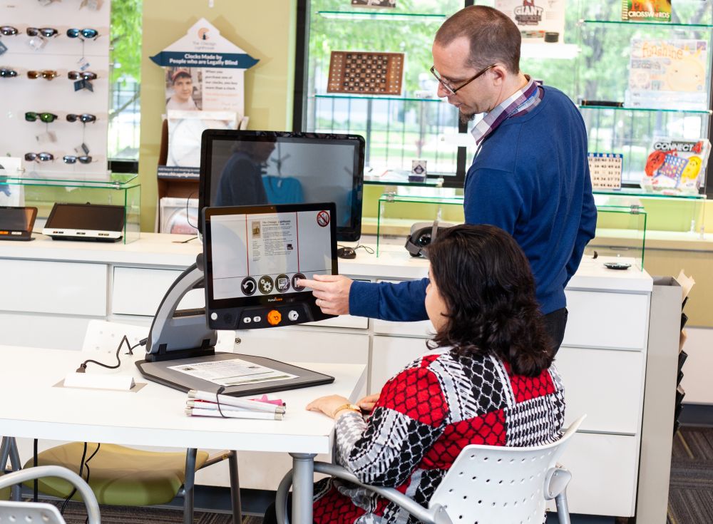 Two people using a large magnifier the size of a small computer screen in the My Tools for Living store. A woman is seated and a man stands next to her pointing
