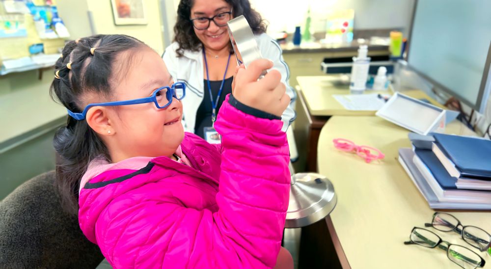 A child with glasses in a pink jacket excitedly examines a pair of eyeglasses in a vision care clinic, with a smiling optometrist observing nearby.
