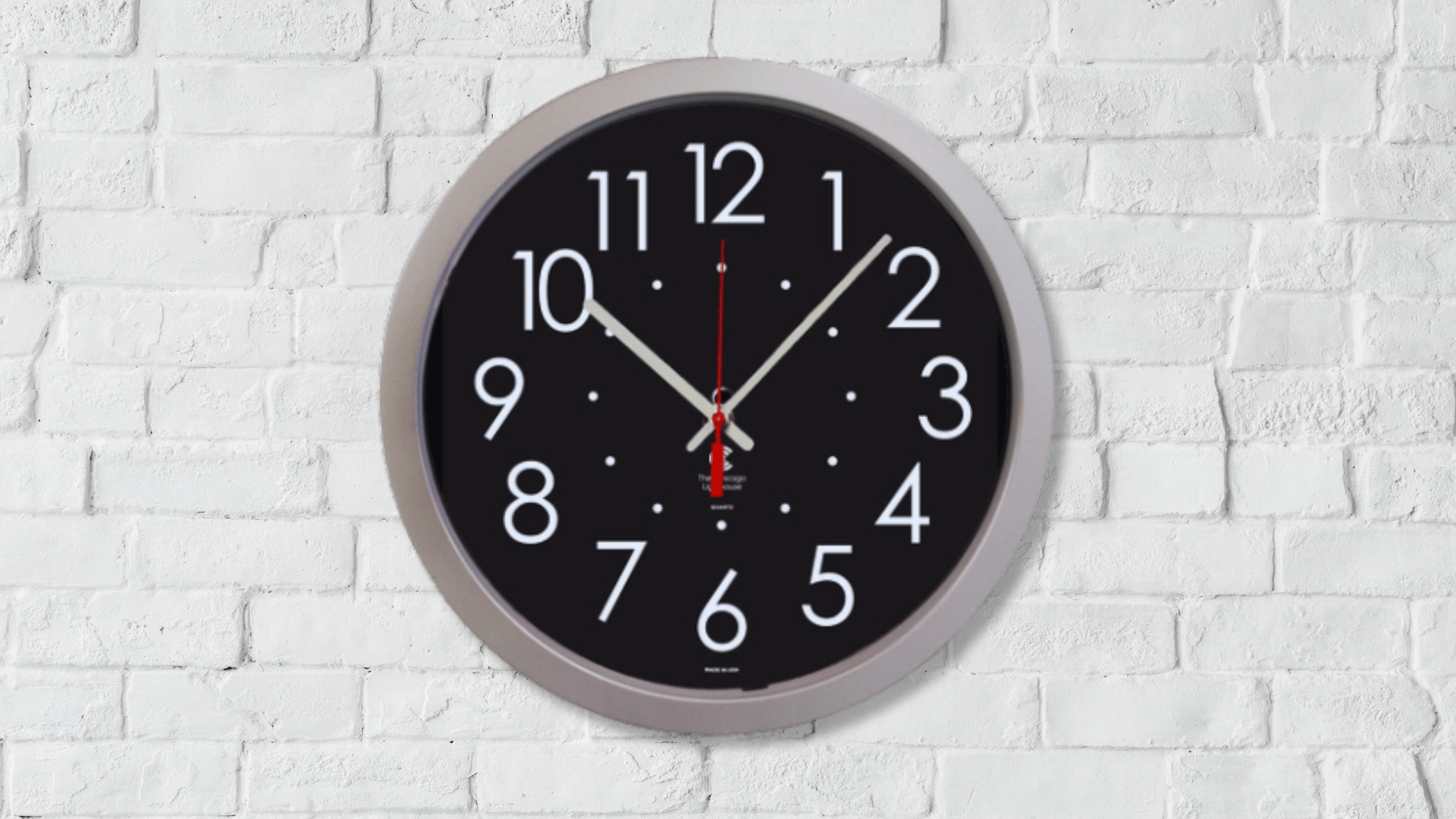 A silver-framed clock with a black background and white numbers hanging on a white brick wall.