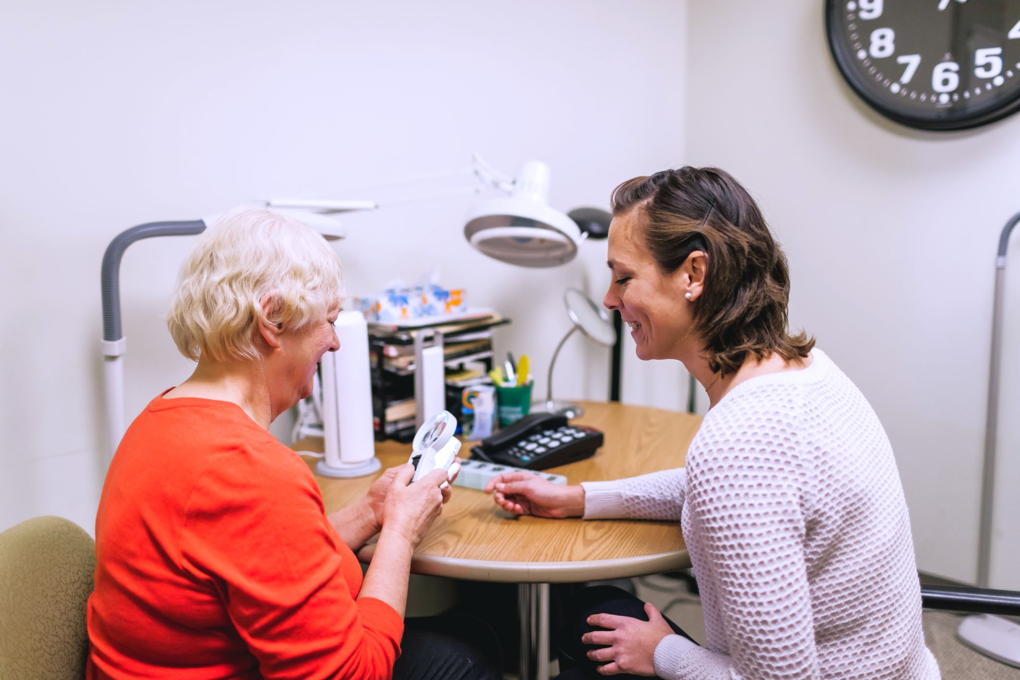 An occupational therapist demonstrating a magnifier to a patient in a clinic office, with a clock and various tools visible in the background.