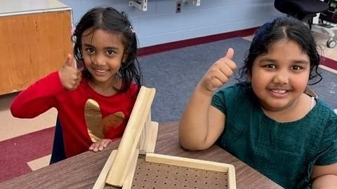 Two young girls in a classroom smiling and holding their right thumbs in the air