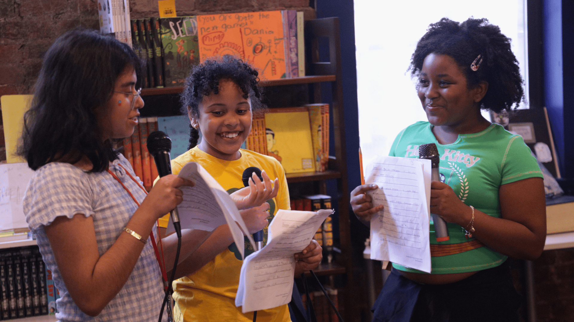 Three 826NYC students perform their play during summer camp. They hold scripts and microphones.