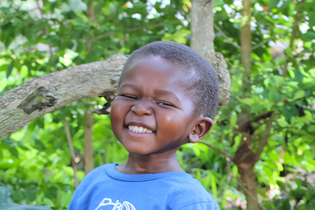 A preschool-aged Ugandan boy smiles so hard that he squints his eyes. He stands under a tree and wears a blue t-shirt.