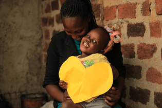A Ugandan girl and her mother are smiling. She is holding up a yellow paper heart.