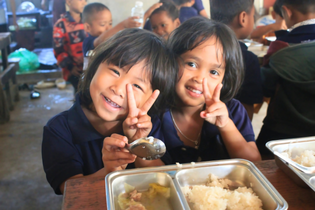 Two Cambodian girls eat their school lunch. They smile at the camera and hold up the peace sign with their fingers.