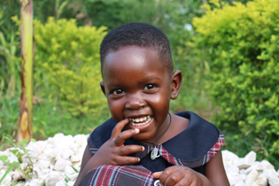 A little girl from Uganda laughs with her hand to her mouth while looking at the camera. She sits in front of trees and wears a black and red dress.