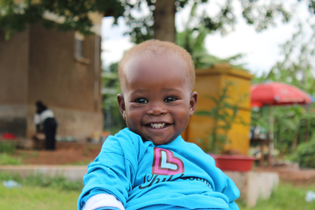 A baby boy from one of LWB's pediatric surgery camps in Uganda. He smiles widely and wears a blue LWB shirt.