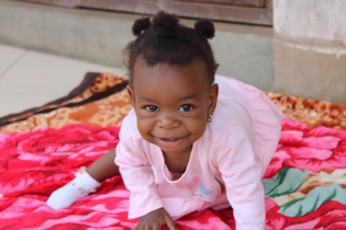 A little baby girl from Uganda looks and smiles at the camera. She crawls on a blanket with large pink roses on it. She is wearing a pink dress and two pigtails