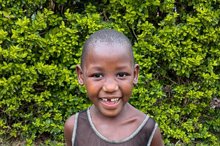 An elementary-aged Ugandan girl with short hair smiles showing her missing front tooth. She stands in front of a hedge.