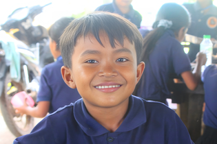 A grade-school-aged boy from Cambodia sits with a smile at a lunch table. He smiles brightly at the camera.