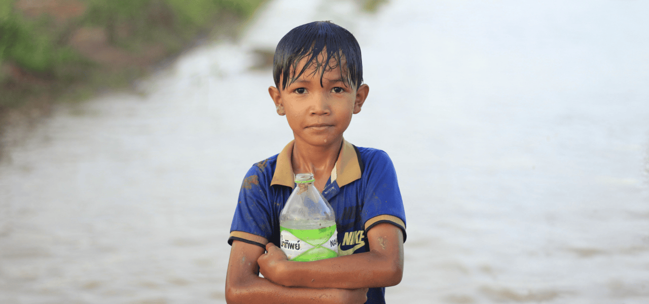 Cambodian boy clutching a plastic water bottle filled with contaminated water from the murky pond behind him.