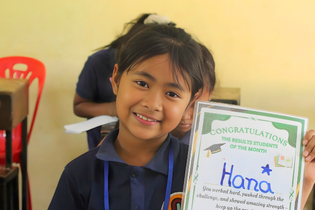 A grade-school-aged girl from Cambodia proudly holds up a certificate. The certificate says, "Hana" and has a star on it.