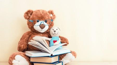 A stuffed brown teddy bear with blue-rimmed glasses reads to a stuffed dog in a blue-striped shirt with a heart on it.