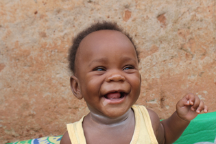 A Ugandan baby boy laughs heartily to the point of drooling. He wears a yellow sleeveless shirt and sits in front of an old wall.
