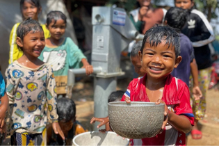 A Cambodian boy holds a large bowl of clean water. He and other children with large smiles on their faces are gathered around their new community well.