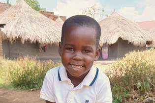 A smiling little Ugandan boy stands in front of houses with straw roofs. He wears a white polo shirt with a blue-rimmed collar.