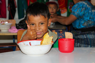 A Guatemalan boy gives a thumbs up for his hot school meal. He sits behind a large bowl of food and a red cup of beverage.