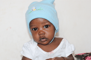 A baby boy in a baby blue knitted beanie and white onesie looks at the camera with curiosity.