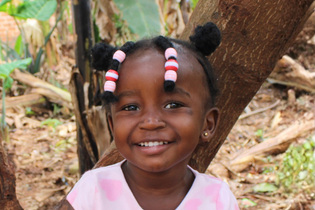A little Ugandan girl with pink, white, and red beads in her hair smiles sweetly at the camera. She wears a pink shirt and sits in front of a tree.