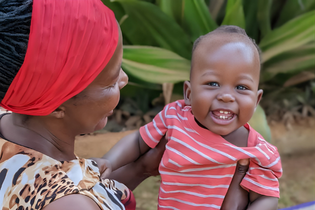 A Ugandan baby boy in a salmon and white striped t-shirt is being held by his foster mother. He smiles brightly with his eyes, revealing his four front teeth.