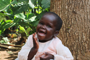 A little girl from Uganda laughs heartily at the camera. She wears a powder pink dress and is in front of a large tree trunk and green bushes.