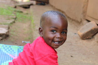 A Ugandan baby boy has his side to the camera but turns his head to smile at it. He wears red and sits on a blue and green mat outdoors.