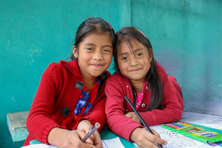 Two school-aged girls taking a pause to smile at the camera while coloring at a table.
