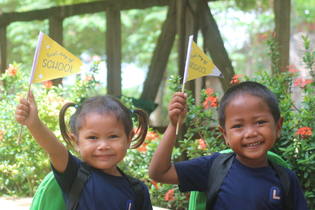 A boy and a girl in LWB's Education Program wave their yellow flags that say "First day of school." They wear their green backpacks and navy school uniform.