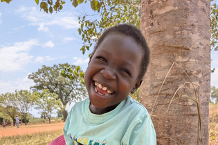 A six-year-old Ugandan girl sits with a big smile in front of a background of beautiful trees and a blue sky. She wears a seafoam colored t-shirt.