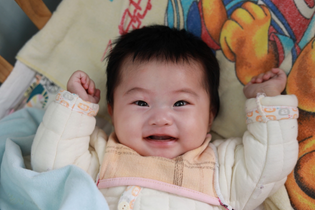 A baby from China laughs at the camera as he lays in a bed full of colorful blankets and pillows. 