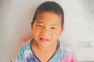 A young boy from China smiles with his tongue out at the camera. He wears a tie-dyed shirt and stands in front of a concrete wall.