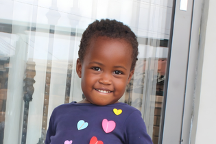 A little Ugandan girl with short hair smiles while biting her lower lip. She wears a dark purple shirt with colorful hearts attached to it.