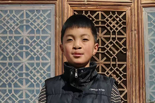 A teenage boy from China with cleft needs stands in front of window shutters with a beautiful geometric pattern. He wears a black winter vest.