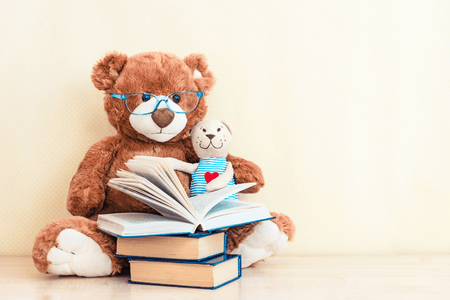 A stuffed brown teddy bear with blue-rimmed glasses reads to a stuffed dog in a blue-striped shirt with a heart on it.