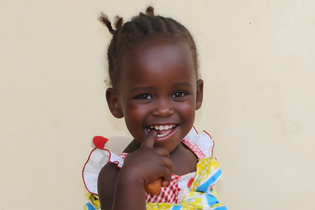A one-year-old girl from Uganda smiles mischievously with a finger in her mouth. She wears a colorful dress and stands in front of a beige wall.