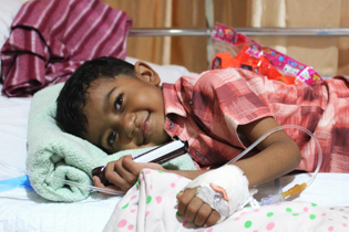 A smiling boy from LWB's Medical Program is resting in bed. He is attached to an IV and holding a granola bar.