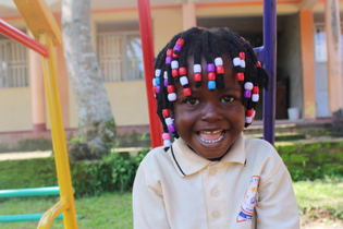 A Ugandan girl of preschool age sits on a play structure with a giant smile. She wears colorful beads in her hair and her beige school uniform.