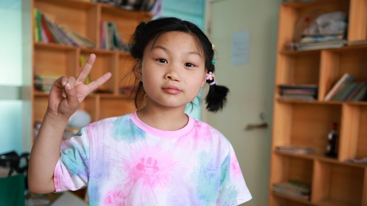 Ruth wears a pink and blue tie-dyed shirt will holding up a peace sign with her right hand.